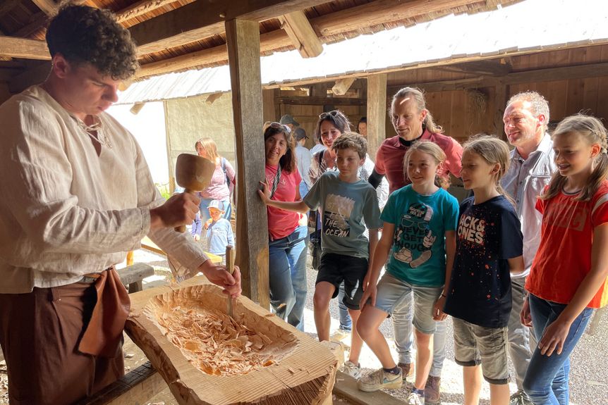 Ein Darsteller in keltischer Gewandung schnitzt Holz und erklärt einer Gruppe neugieriger Kinder das Handwerk.