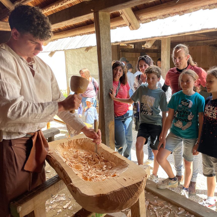 Ein Darsteller in keltischer Gewandung schnitzt Holz und erklärt einer Gruppe neugieriger Kinder das Handwerk.