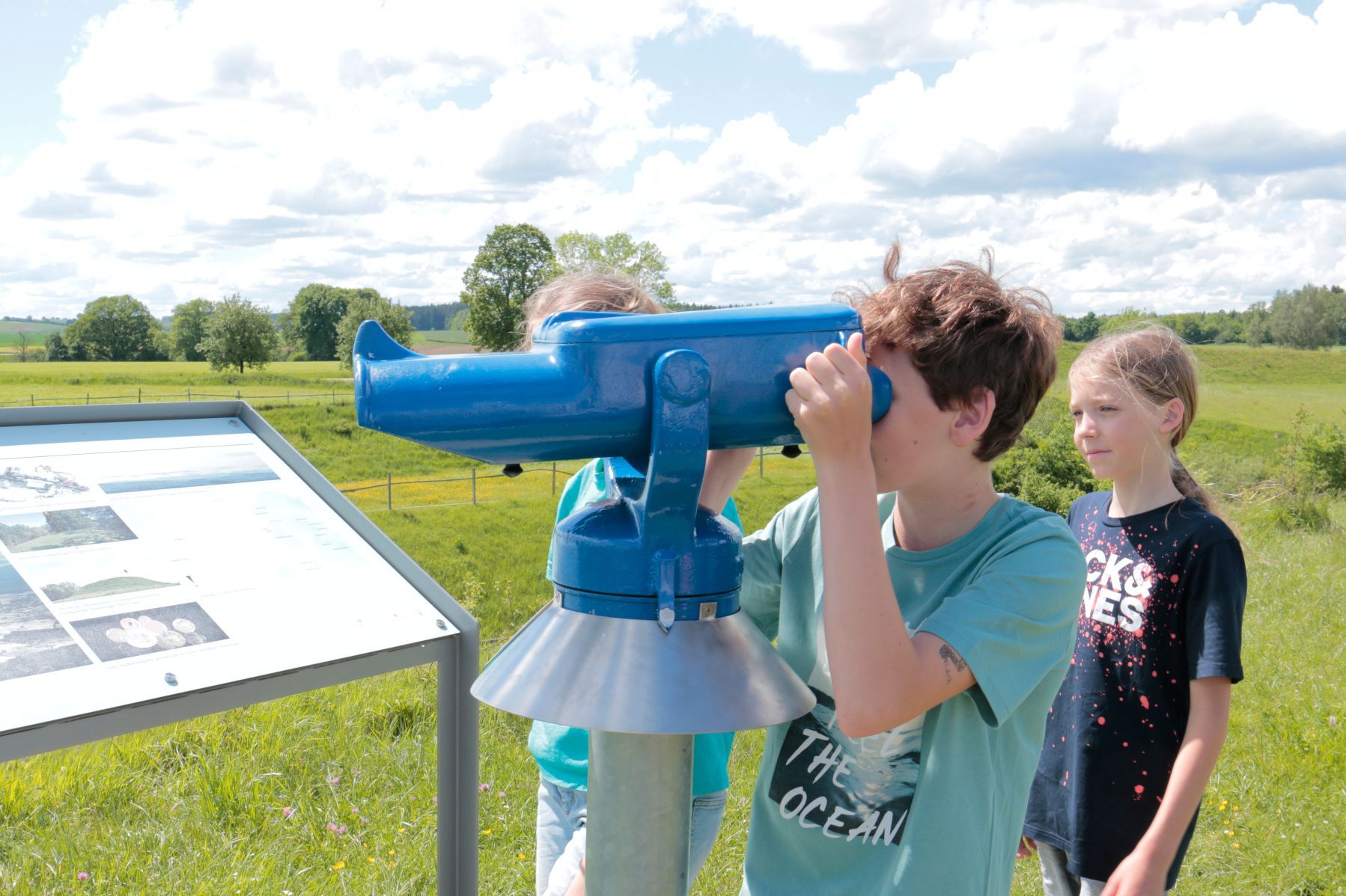 Kinder betrachten durch ein Fernglas die Umgebung der Heuneburg – Stadt Pyrene. Im Hintergrund grüne Wiesen und Informationstafel zur keltischen Höhensiedlung