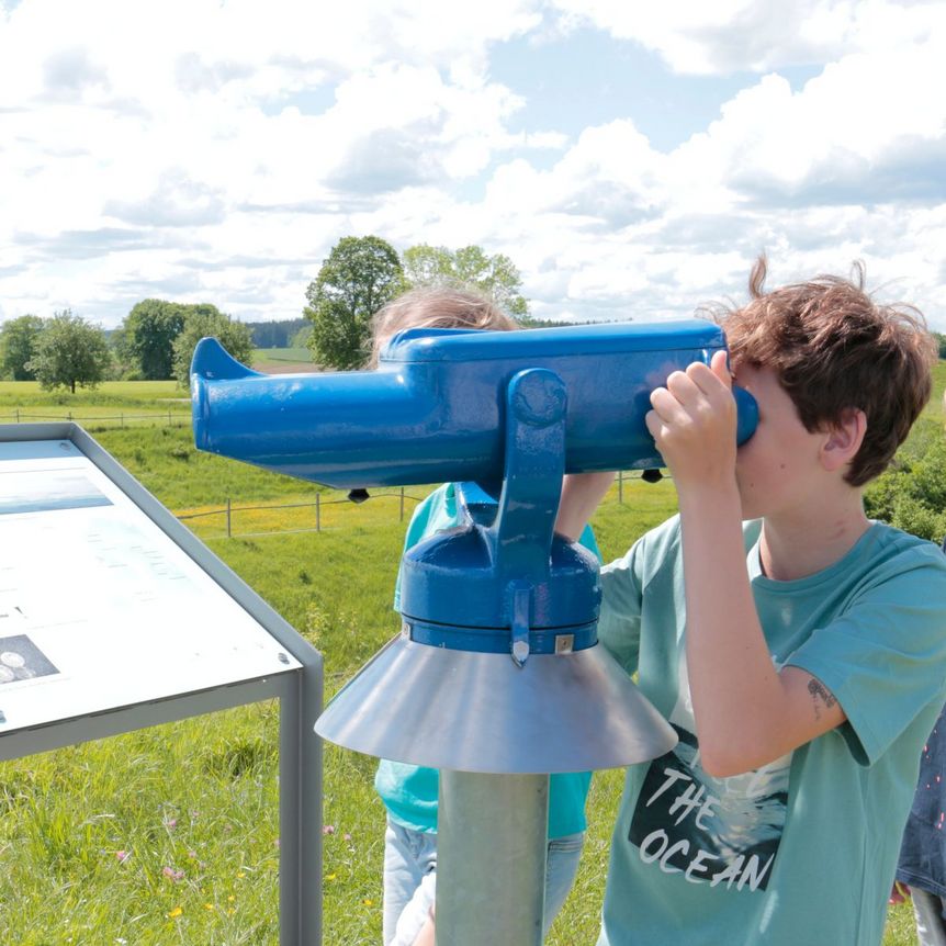 Kinder betrachten durch ein Fernglas die Umgebung der Heuneburg – Stadt Pyrene. Im Hintergrund grüne Wiesen und Informationstafel zur keltischen Höhensiedlung